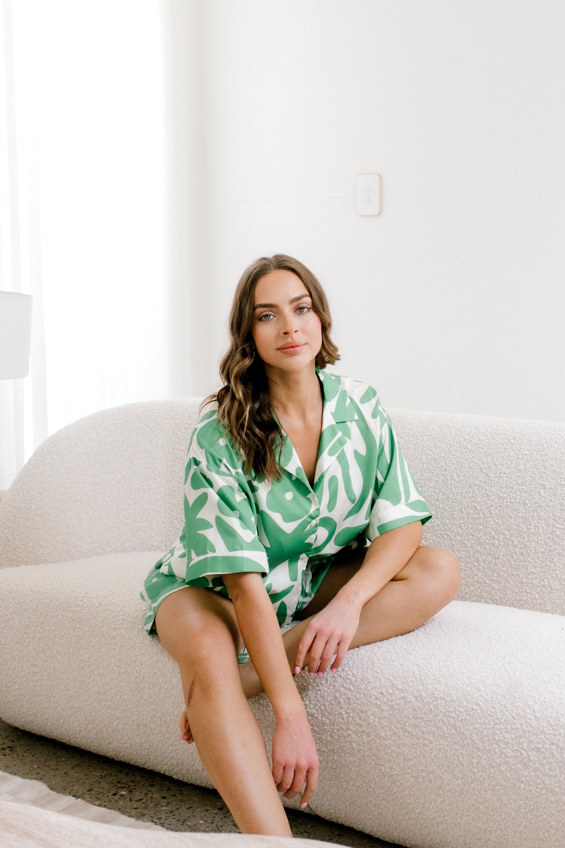 Woman in a green patterned pyjamas sitting on a white couch in a bright room.